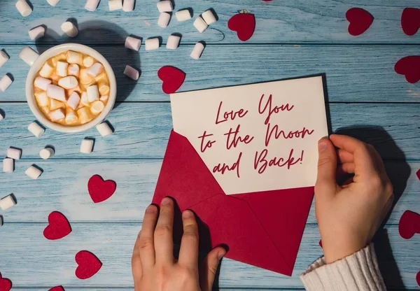 LOVE YOU TO THE MOON AND BACK text on valentine card inscription positive quote, motivation and inspiration phrase. Female hands holding valentine card red envelope with white cup of coffee and marshmallows on wooden blue background. Romantic Small h