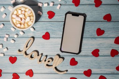Greeting or invitation white screen on mobile phone mock up with white cup of coffee and marshmallows on wooden background. Romantic Small hearts Valentine day. Blank card copy space for your text. Holiday morning. Top view, flat lay minimalist aesth