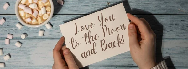 LOVE YOU TO THE MOON AND BACK text on valentine card inscription positive quote phrase. Banner Female hands holding greeting card with white cup of coffee and marshmallows on wooden blue background