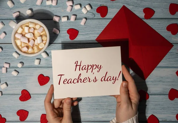 HAPPY TEACHERS DAY text Female hands holding valentine card red envelope with white cup of coffee and marshmallows on wooden blue background. Holiday morning. Top view, flat lay minimalist branding