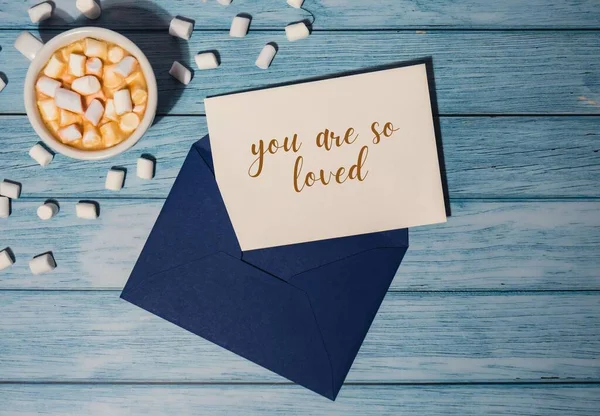 YOU ARE SO LOVED text on valentine postcard inscription positive quote phrase Greeting card blue envelope with white cup of coffee and marshmallows on wooden blue background holiday. Top view, flat