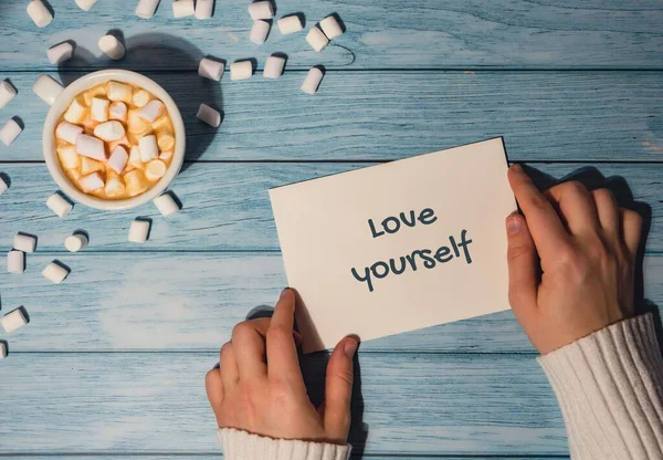 LOVE YOURSELF text on valentine card. Female hands holding greeting card with white cup of coffee and marshmallows on wooden blue background. holiday. Top view, flat lay minimalist branding concept