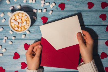 Female hands holding empty valentine card mock up red envelope with white cup of coffee and marshmallows on wooden blue background. Romantic Small hearts Valentine day. Blank paper card copy space for