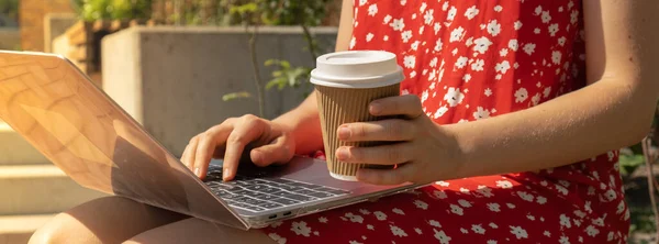 Unrecognizable Young woman in red dress drinking Take away coffee in craft recycling paper cup with laptop. Freelancer\'s place of work. Study and work online. Remote business education. View webinar. E-learning Workstation on wooden bench. Mockup Cof