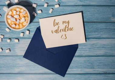 BE MY VALENTINE text Greeting card blue envelope with white cup of coffee and marshmallows on wooden blue background holiday. Top view, flat lay minimalist