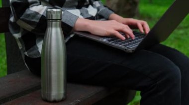 Su şişesi. Ahşap bankta yeniden kullanılabilir çelik termo su şişesi. Sürdürülebilir yaşam tarzı. Plastiksiz sıfır atıksız yaşam. Yeşil çevre korumasına geçin. Dizüstü bilgisayarla çalışan bir kadın öğrenci.