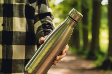 Close-up of Unrecognizable female hand holding Water bottle. Reusable steel thermo water bottle in park. Sustainable lifestyle. Plastic free zero waste free living. Go green Environment protection. Woman drinking water in park 