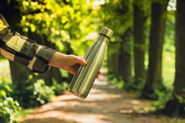 Close-up of Unrecognizable female hand holding Water bottle. Reusable steel thermo water bottle in park. Sustainable lifestyle. Plastic free zero waste free living. Go green Environment protection. Woman drinking water in park 