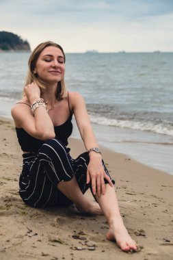 Young woman siting on blurred beachside background. Attractive female enjoying the sea shore. travel and active lifestyle concept. Springtime. Relaxation, youth, love, lifestyle solitude with nature. Wellness wellbeing mental health inner peace Slow 