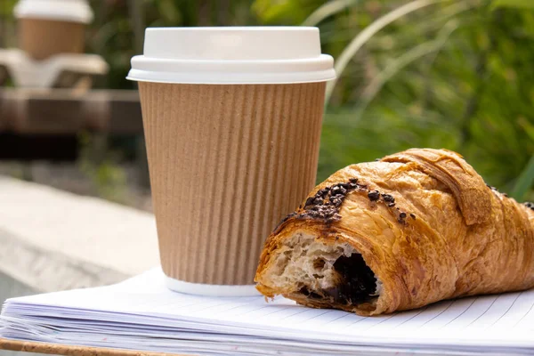 Fresh baked chocolate croissant on paper cup with lid for tea to go. Empty paper notebook. Freelancer\'s place of work. Study and work online. Remote business education. Making plans for next week year. Being mindful reducing stress and slowing-down.