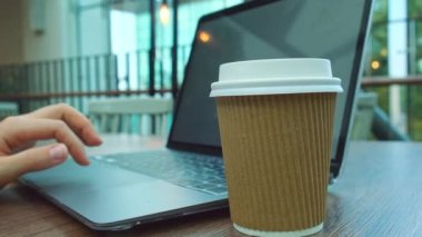 Tanınmayan genç bir kadın alışveriş merkezindeki ahşap masada modern bir dizüstü bilgisayarda çalışıyor. Kağıt bardaktan kahve içmek. Eş zamanlı çalışma alanında klavyede yazan Öğrenci Çalışan Kadın Elleri