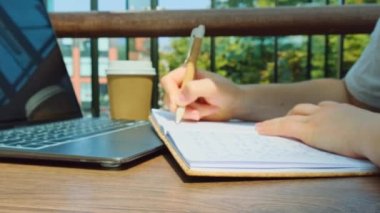 Alışveriş merkezinin yemek bölümündeki ahşap masada tanınmayan genç bir kadın ders çalışıyor. Kağıt bardaktan kahve içmek. Öğrenci ev ödevi yapıyor. Kadın elleri deftere yazıyor. Minnettarlık günlüğü yansıması.