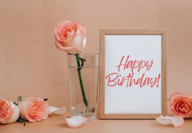 Mockup Wooden frame with text HAPPY BIRTHDAY on blank and delicate pink roses on beige background. Minimal trendy composition. Romantic pastel pink rose flowers. Neutral earth tones. Greeting card