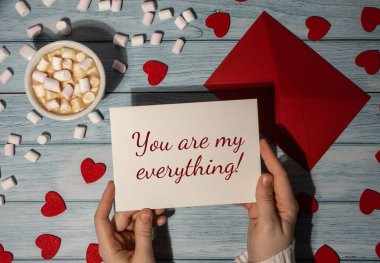 YOU ARE MY EVERYTHING valentine text Female hands holding valentine card red envelope with white cup of coffee and marshmallows on wooden blue background. Romantic Small hearts Valentine day. Holiday