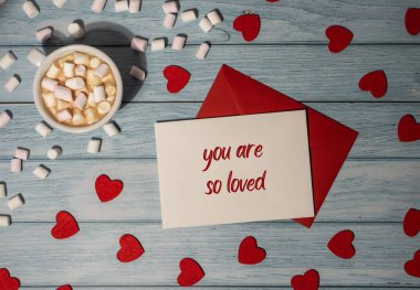 YOU ARE SO LOVED text on valentine postcard inscription positive quote phrase Greeting card with red envelope with white cup of coffee and marshmallows on wooden background. Romantic Small hearts