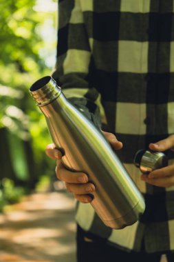 Tanımlanamayan bir kadının su şişesini tuttuğu yakın plan. Parkta yeniden kullanılabilir çelik termo su şişesi. Sürdürülebilir yaşam tarzı. Plastiksiz sıfır atıksız yaşam. Yeşil Çevre Koruma 'ya git