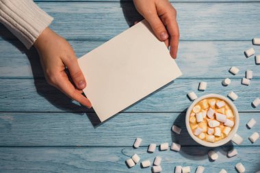 Female hands holding empty greeting or invitation card mock up with white cup of coffee and marshmallows on wooden blue background. Blank paper copy space for your text. Valentines day holiday. Top