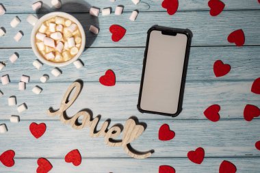 Greeting or invitation white screen on mobile phone mock up with white cup of coffee and marshmallows on wooden background. Romantic Small hearts Valentine day. Blank card copy space for your text