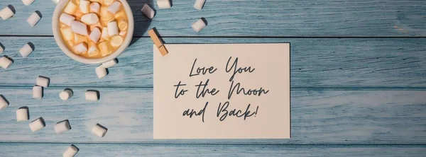 LOVE YOU TO THE MOON AND BACK text on valentine card inscription positive quote phrase. Greeting card with white cup of coffee and marshmallows on wooden blue background. holiday concept. Top view