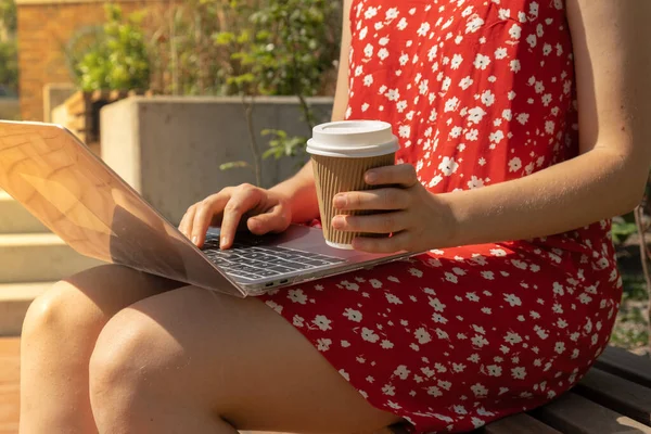 Unrecognizable Young woman in red dress drinking Take away coffee in craft recycling paper cup with laptop. Freelancers place of work. Study and work online. Remote business education. View webinar