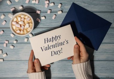 HAPPY VALENTINES DAY text Female hands holding greeting card blue envelope with white cup of coffee and marshmallows on wooden blue background. Top view, flat lay minimalist branding concept