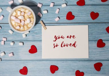 YOU ARE SO LOVED text on valentine postcard inscription positive quote phrase Greeting card with red envelope with white cup of coffee and marshmallows on wooden background. Romantic Small hearts