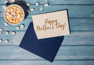 HAPPY MOTHERS DAY text on card inscription positive quote phrase Greeting card blue envelope with white cup of coffee and marshmallows on wooden blue background holiday. Top view, flat lay minimalist