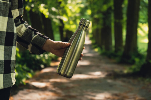Close-up of Unrecognizable female hand holding Water bottle. Reusable steel thermo water bottle in park. Sustainable lifestyle. Plastic free zero waste free living. Go green Environment protection