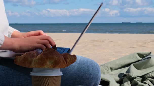 海の景色の隣のビーチでノートパソコンで働く若い女性 コーヒーを飲み クロワッサンを食べる 学生勉強は暖かい秋の海岸の屋外で動作します フリーランス — ストック動画