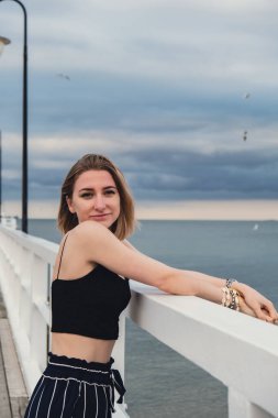 Young woman standing on wooden pier blurred beachside background. Attractive female enjoying the sea shore travel and active lifestyle concept. Springtime. Wellness wellbeing mental health inner peace
