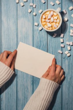 Female hands holding empty greeting or invitation card mock up with white cup of coffee and marshmallows on wooden blue background. Blank paper copy space for your text. Valentines day holiday. Top