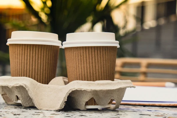 Two paper cups with lid for tea to go. Coffee take away on the table. Take-out coffees with brown paper cup holder. Brown safety cardboard collars. Take away box for cups. Cardboard tray