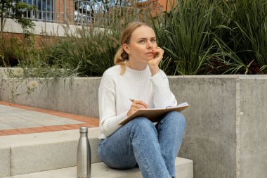 Young student study with notebook in park. Drinking water, hot tea or coffee from reusable metal bottle. Writing gratitude journal self reflection self discovery Outdoors warm autumn seashore.  clipart