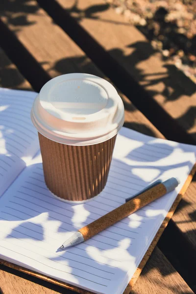 Eco recycling paper cup with coffee or tea on kraft paper with empty paper notebook on wooden bench. Concept of study work outdoors. Take away coffee to go. Copy space for text. Disposable Cardboard