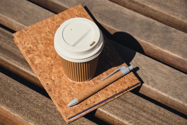 Eco recycling paper cup with coffee or tea on kraft paper with empty paper notebook on wooden bench. Concept of study work outdoors. Take away coffee to go. Copy space for text. Disposable Cardboard