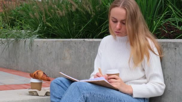 公園のノート付きの若い学生の勉強 コーヒーを飲み クロワッサンを食べる 感謝日記自己反省自己発見を書く屋外暖かい秋の海岸 — ストック動画