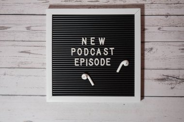 Yeni PODCAST EPISODE yazılı duyuru panosu ahşap masada kablosuz kulaklıklarla. Podcast konsepti. Radyodaki ses iyileştirme sağlık alışkanlıkları.