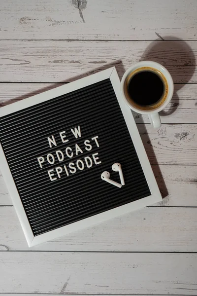 Tahta masada üzerinde yeni PODCAST EPISODE yazılı beyaz kahve fincanı kablosuz kulaklıkla duyuru panosu. Podcast konsepti. Radyodaki ses iyileştirme sağlık alışkanlıkları.