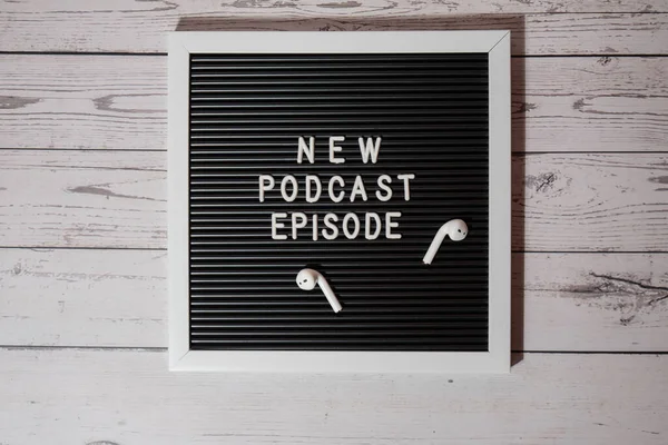 Yeni PODCAST EPISODE yazılı duyuru panosu ahşap masada kablosuz kulaklıklarla. Podcast konsepti. Radyodaki ses iyileştirme sağlık alışkanlıkları.