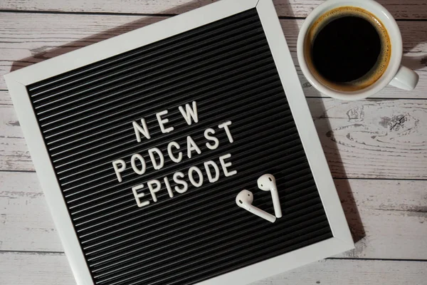 Tahta masada üzerinde yeni PODCAST EPISODE yazılı beyaz kahve fincanı kablosuz kulaklıkla duyuru panosu. Podcast konsepti. Radyodaki ses iyileştirme sağlık alışkanlıkları.