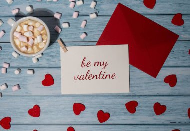 BE MY VALENTINE text Greeting card with red envelope with white cup of coffee and marshmallows on wooden background. Romantic Small hearts Valentine day. Holiday morning. Top view, flat lay minimalist