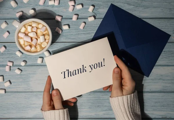 感謝のはがきの碑文の肯定的な引用句のありがとうテキスト女性の手は木製の青の背景にコーヒーとマシュマロの白いカップとグリーティングカードブルー封筒を保持します トップ表示 — ストック写真