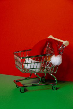 Santa hat in shopping trolley on isometric red green background. Online shopping holiday concept Copy space for your text. Mock up for advertisement. Holiday Christmas composition. Minimalistic