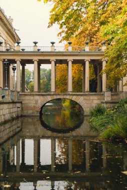 Baths classicist Palace on the Isle in Lazienki Park touristic place in Warsaw. Lazienki Royal Baths Park, Warsaw Poland. Colorful Autumn Foliage and Mirror Reflection on the Lake. Yellow leaves