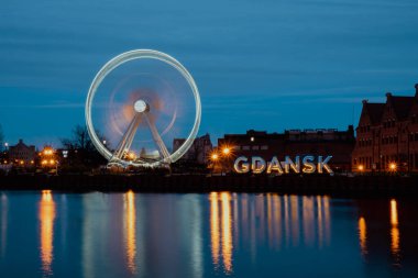 Gdansk Polonya Dönme Dolabı, eski Gdansk kasabasında akşamüstü Avrupa nehri suyunda alacakaranlık yansıması. Uzun pozlama fotoğrafı. Şehir manzarası aydınlatılmış eğlence parkı ve sokak güzelliğiyle