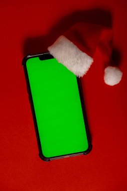 Mobile phone dressed in Santa-Claus red-white hat with chroma key screen against red background. Concept for Christmas or New Years holidays. Blank cell phone. Digital gadget, technology copy space