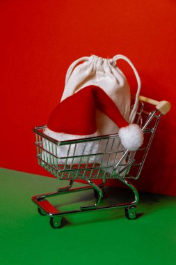 Santa hat in shopping trolley on isometric red green background. Online shopping holiday concept Copy space for your text. Mock up for advertisement. Holiday Christmas composition. Minimalistic