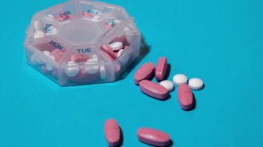Düzenleyicinin haftalık görüntülerini mavi arkaplanda metnin için kopyalama alanı ile yakınlaştır. Tıbbi hap kutusunun kapanışı, günlük ilaç dozları, beyaz pembe haplar ve kapsüllerle. Günlük