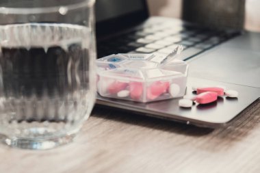 Organizer weekly shots with glass of water on workplace background and laptop Closeup of medical pill box with doses of tablets for daily take medicine with white pink drugs and capsules. Medication