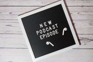 Yeni PODCAST EPISODE yazılı duyuru panosu ahşap masada kablosuz kulaklıklarla. Podcast konsepti. Radyodaki ses iyileştirme sağlık alışkanlıkları.